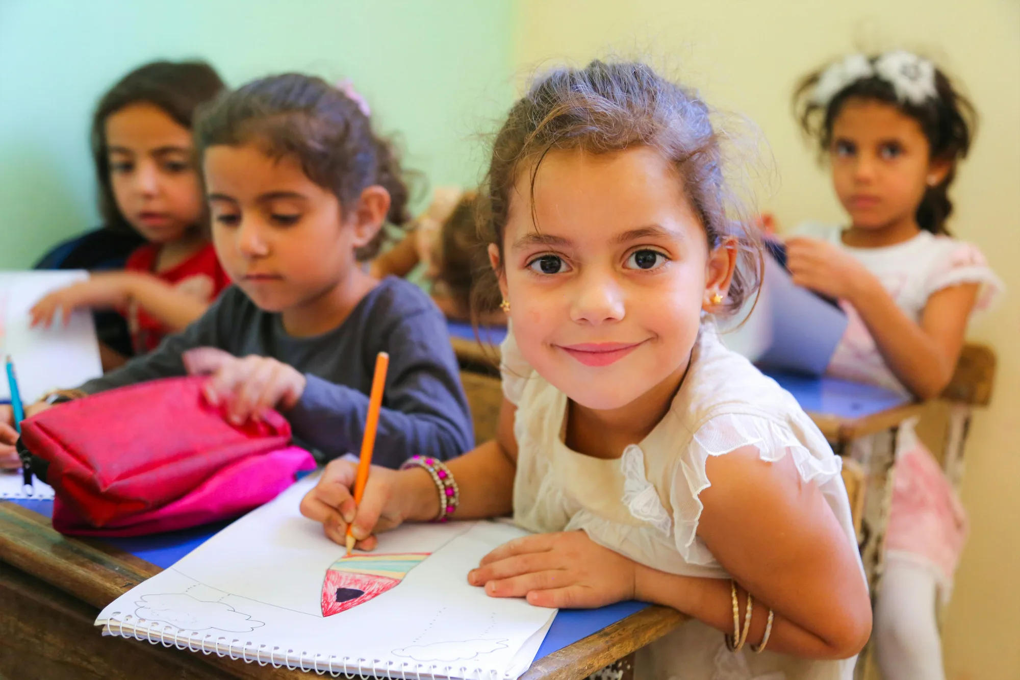 Smiling schoolgirl drawing at her desk with classmates in a classroom